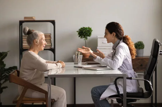 Medicare for older people. Friendly hispanic woman gp family therapist consult elderly female patient at meeting at hospital cabinet. Old lady retiree visit doc listen to recommendation for treatment